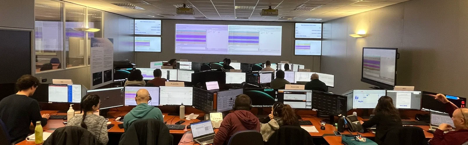 Sala de control con varias personas de espaldas ante filas de monitores y una gran pantalla al fondo, en el centro de operaciones del rover de Turín