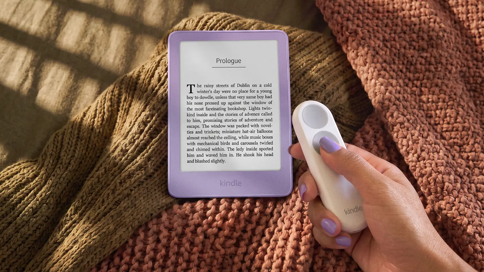 Una mano sujeta el mando Kindle Click junto a un Kindle básico morado con una novela en pantalla, sobre mantas de punto