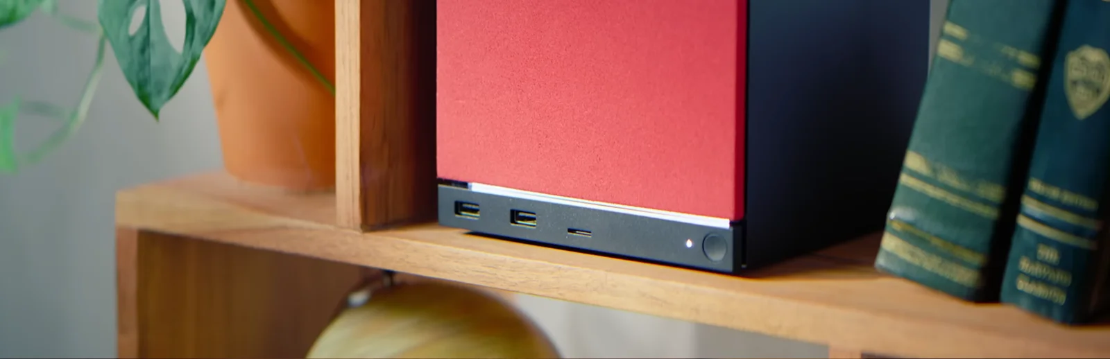 Steam Machine con la placa frontal de tela roja en una estantería de madera, junto a una planta y unos libros