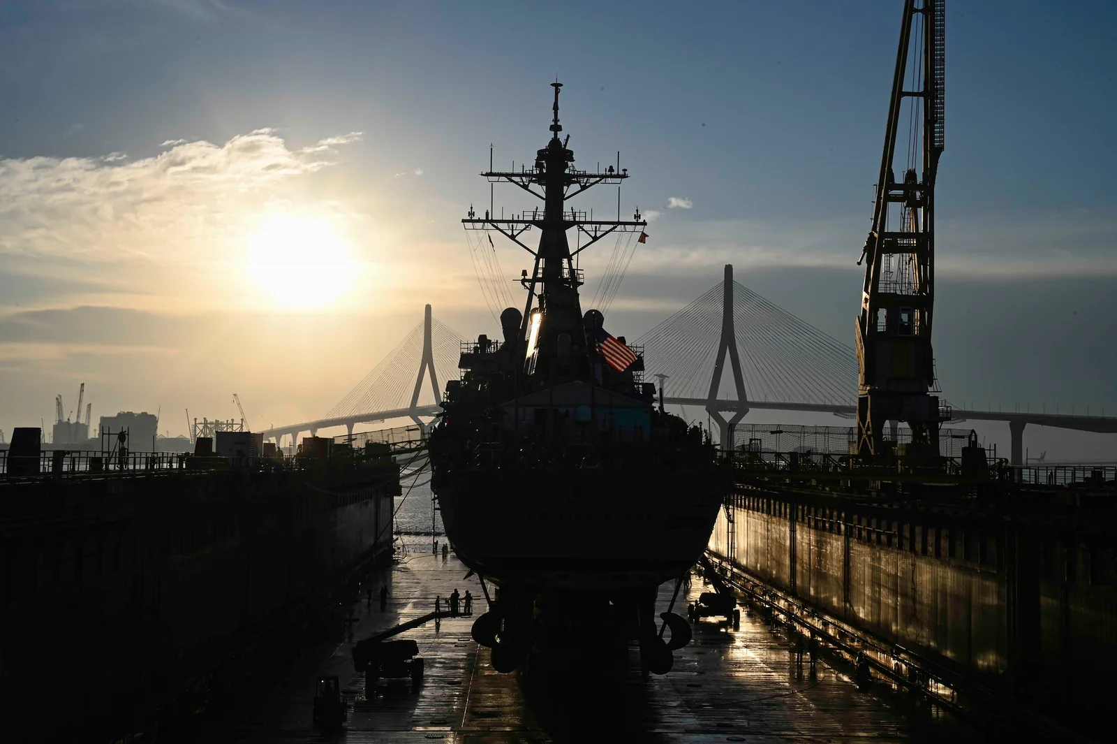 Silueta del mástil del destructor USS Arleigh Burke en el dique de Navantia en Cádiz, con la bandera de Estados Unidos, una grúa del astillero y el puente de la Constitución de 1812 al fondo, al atardecer
